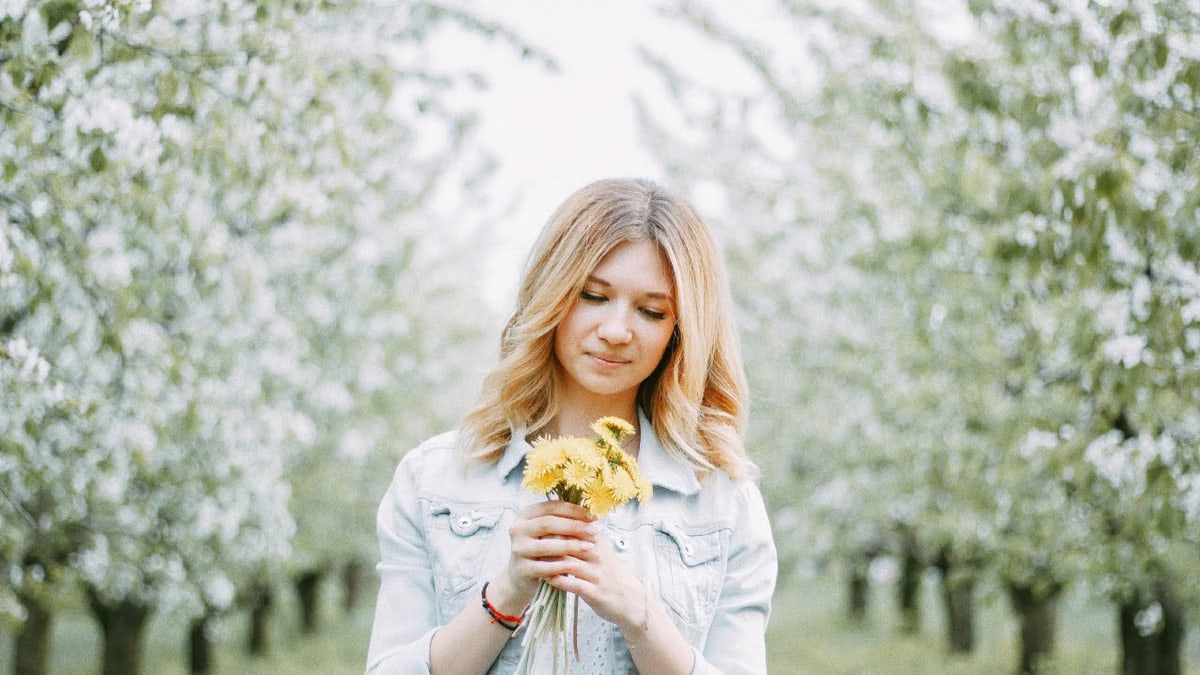 A young woman with blond hair holds dandelions in a blooming orchard, signifying springtime beauty.